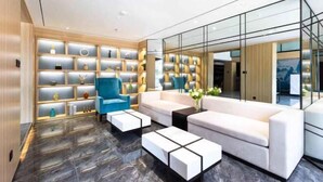 Un vestíbulo moderno con suelo de mármol, un sillón azul, un sofá blanco y un muro de cristal que refleja el interior.