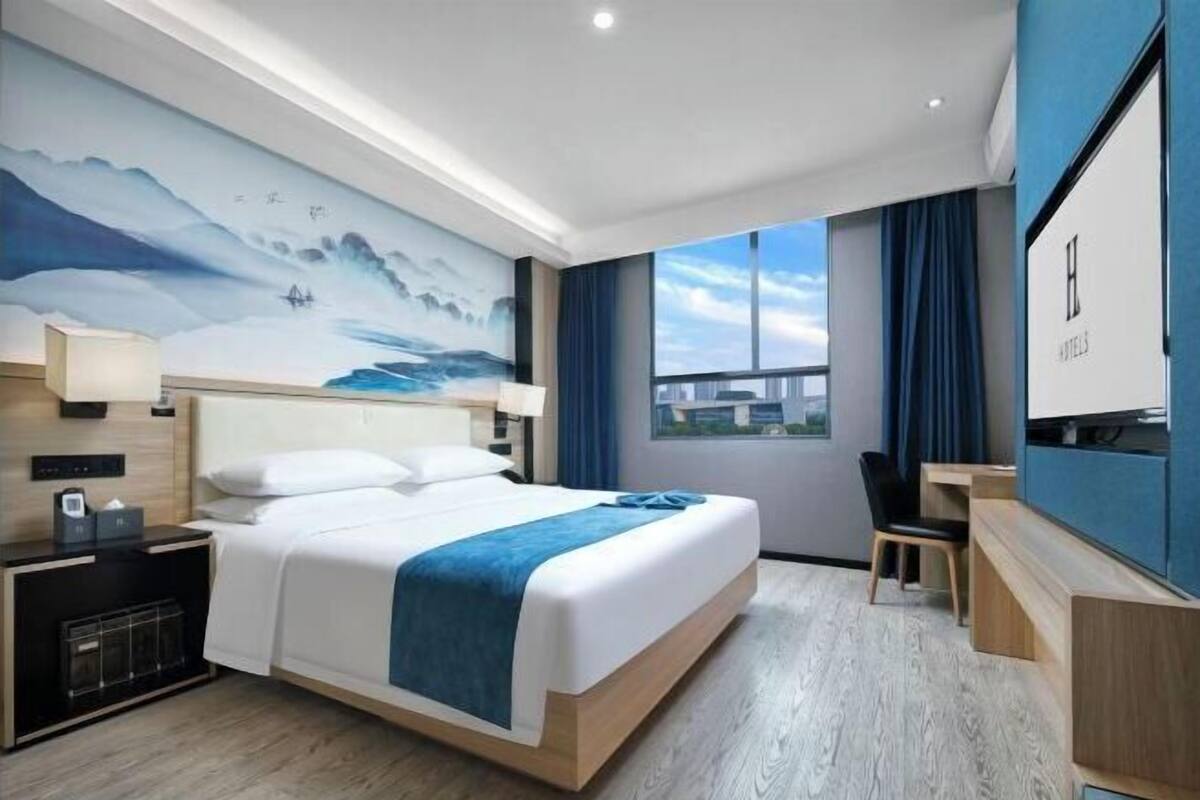 A modern hotel room with a large bed, a desk, a chair, a TV, and a painting on the wall.