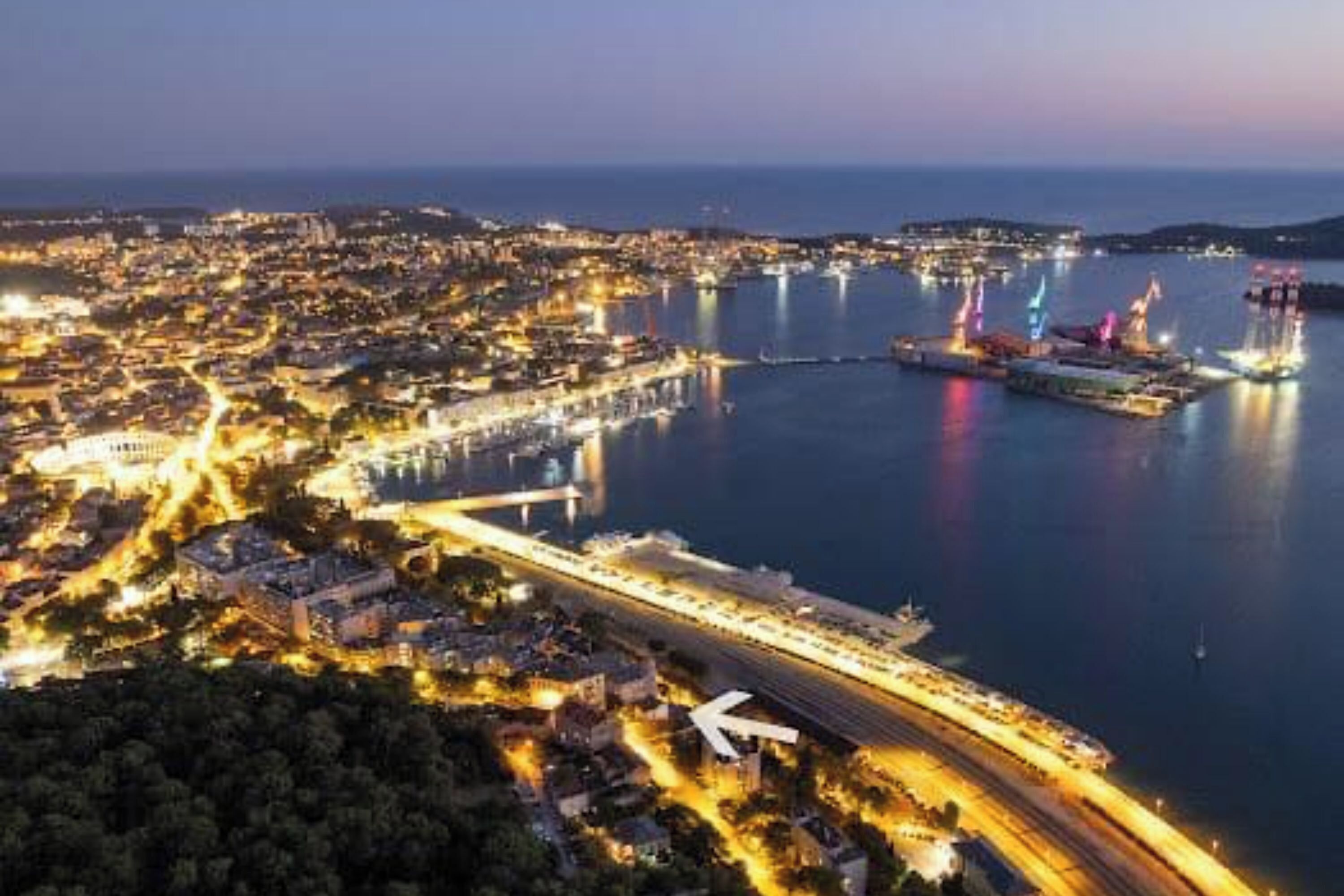 An aerial view of a coastal city at dusk with a prominent bridge and a large cruise ship docked at the port.