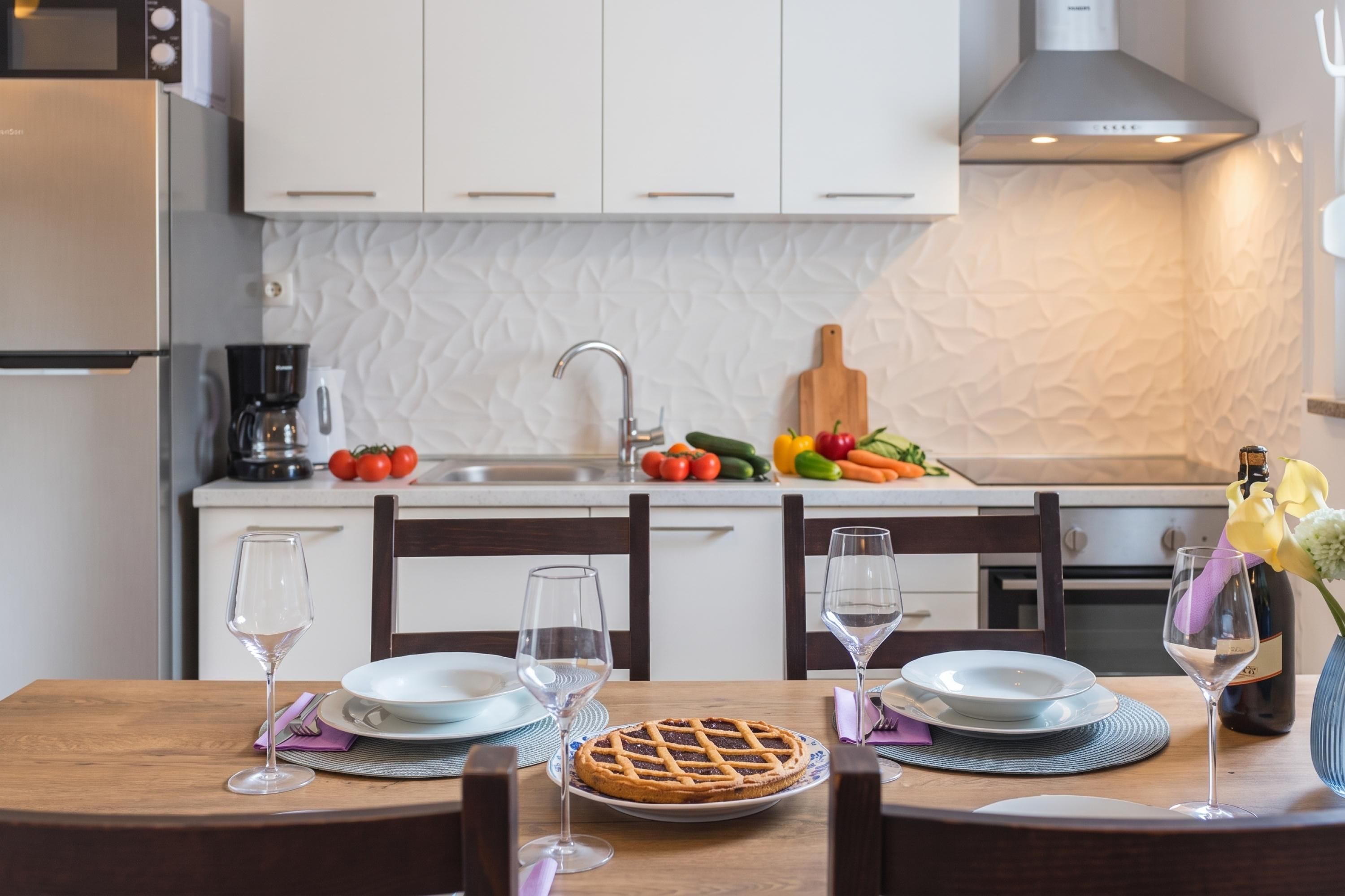 Une cuisine moderne avec des meubles blancs, un réfrigérateur en acier inoxydable, une machine à café et un panier de fruits posé sur le plan de travail.