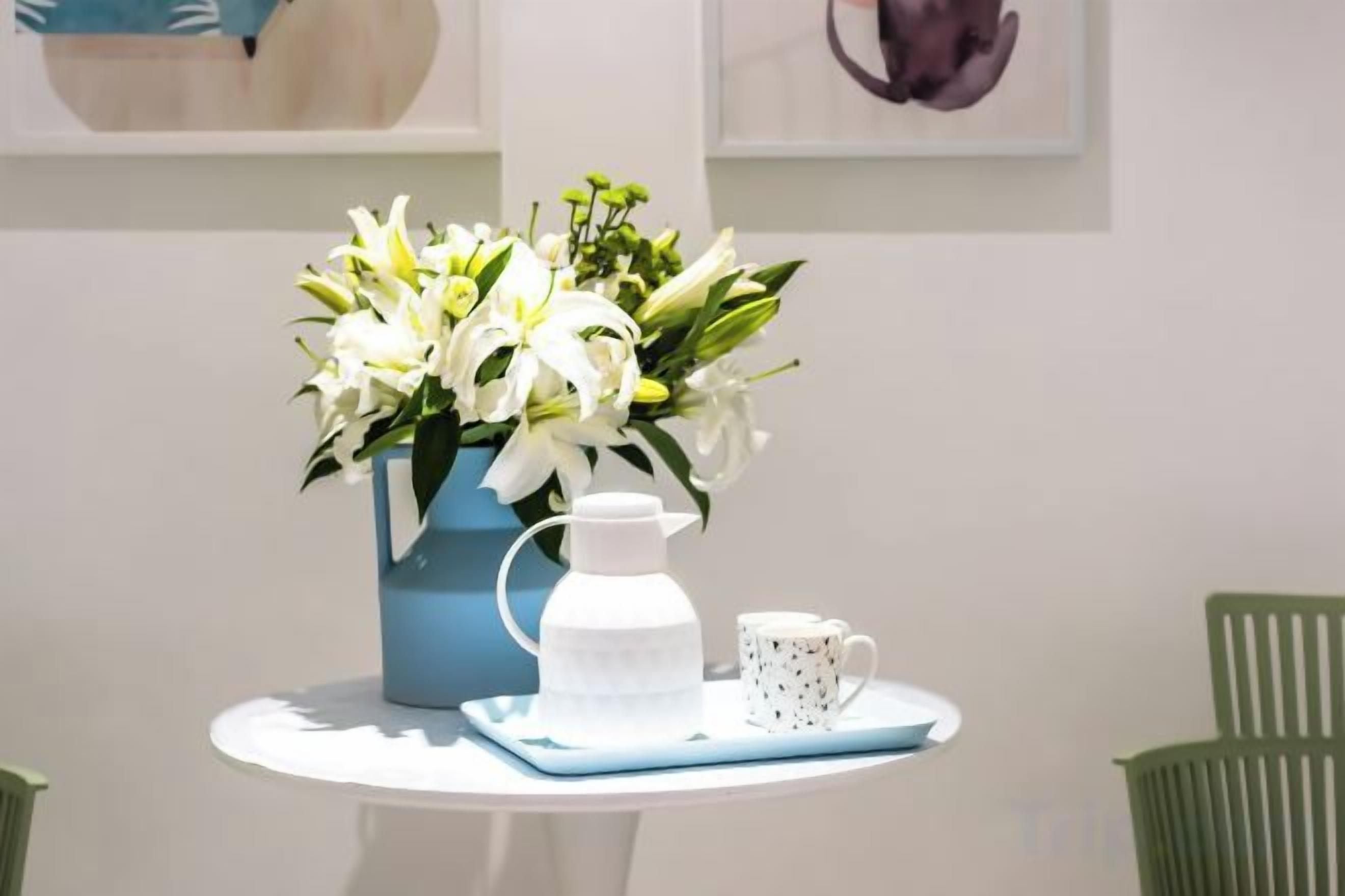 A table with a blue vase of white flowers, a white pitcher, and a white mug on a white table.