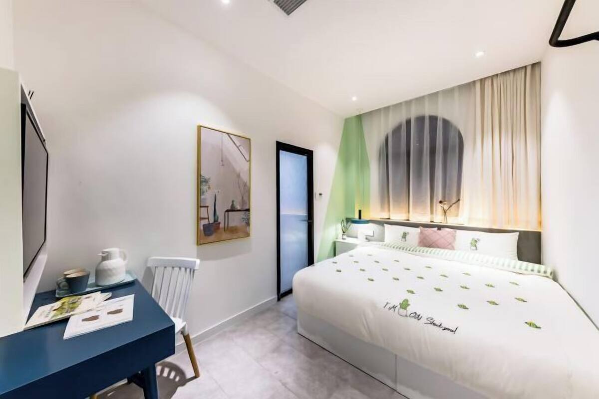 A modern hotel room with a large bed, a desk with a chair, a radiator, and a painting on the wall.