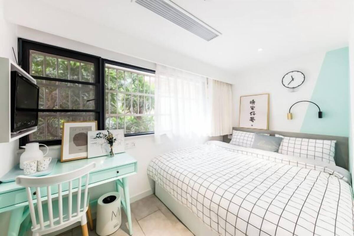 A bedroom with a bed, a desk, a chair, a window with curtains, a clock, and a wall-mounted lamp.