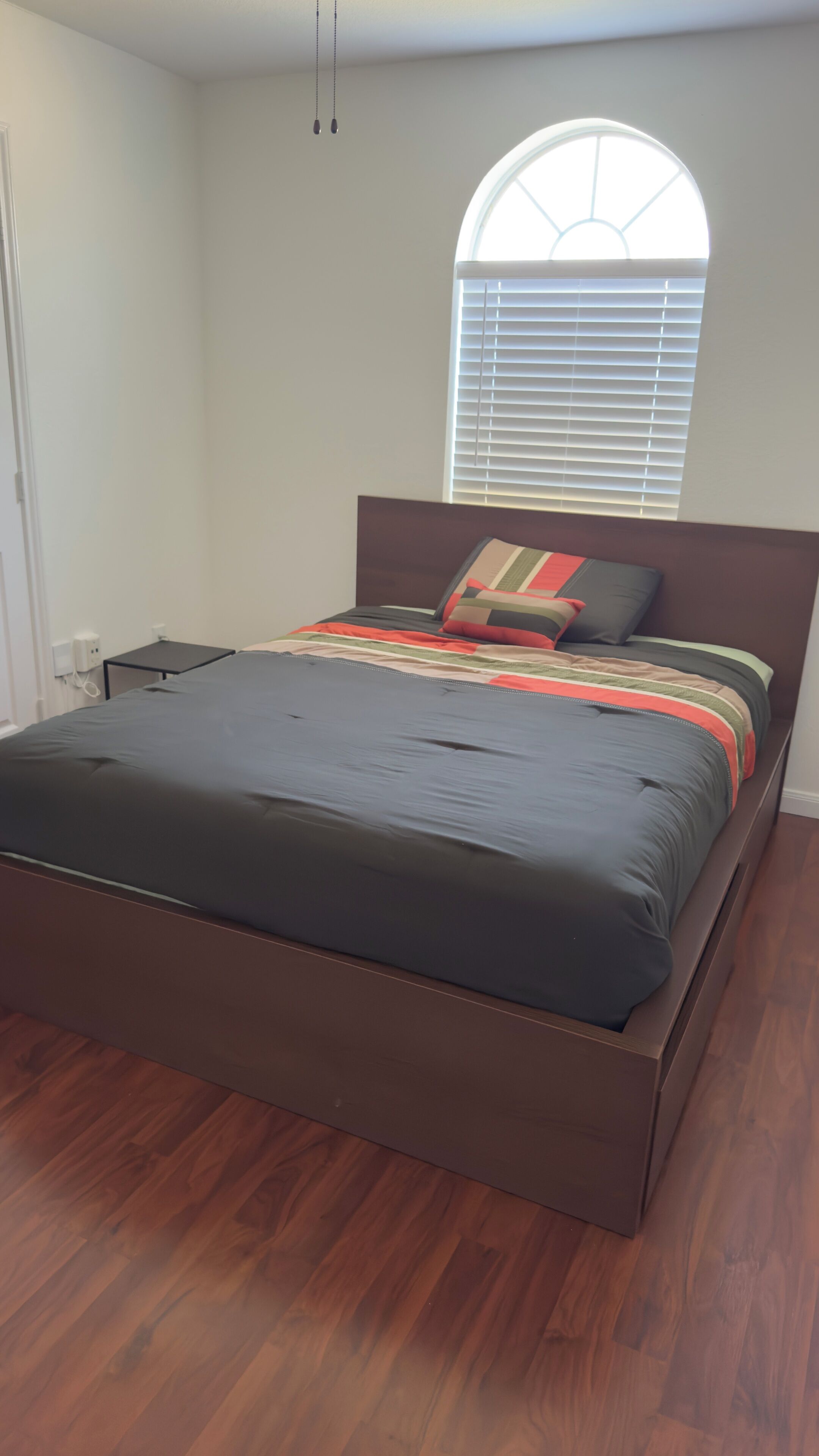 A bedroom with a bed, a nightstand, a window with blinds, and a hanging light fixture.