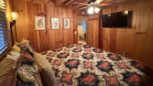 A cozy room with wooden walls, a large bed with a patterned blanket, a ceiling fan, and a TV mounted on the wall. - Cabin, Hot Tub, Forest View, Close to Town (West Jefferson)