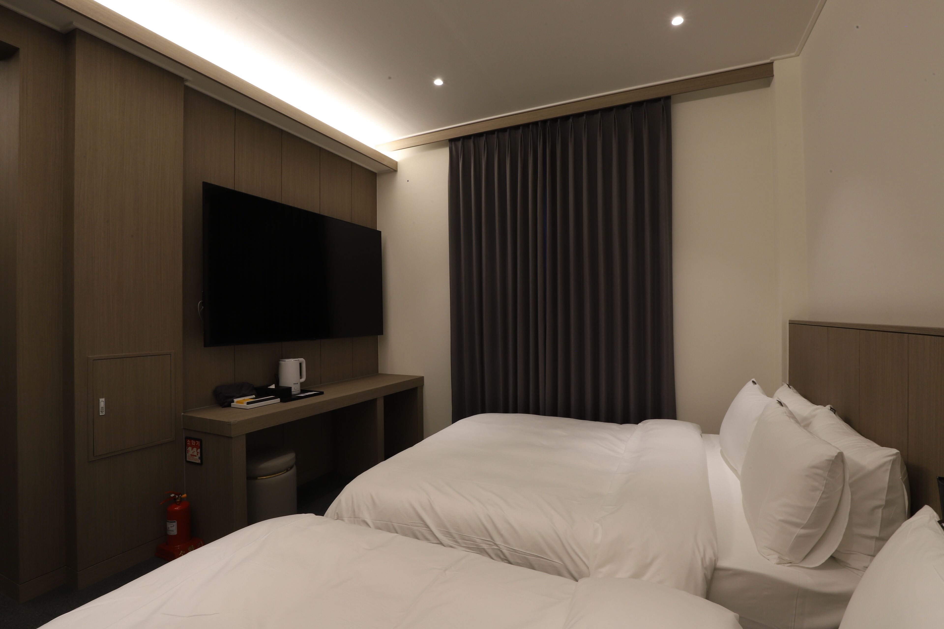 A hotel room with two beds, a flat-screen TV mounted on the wall, a small desk with a coffee maker, and a fire extinguisher.