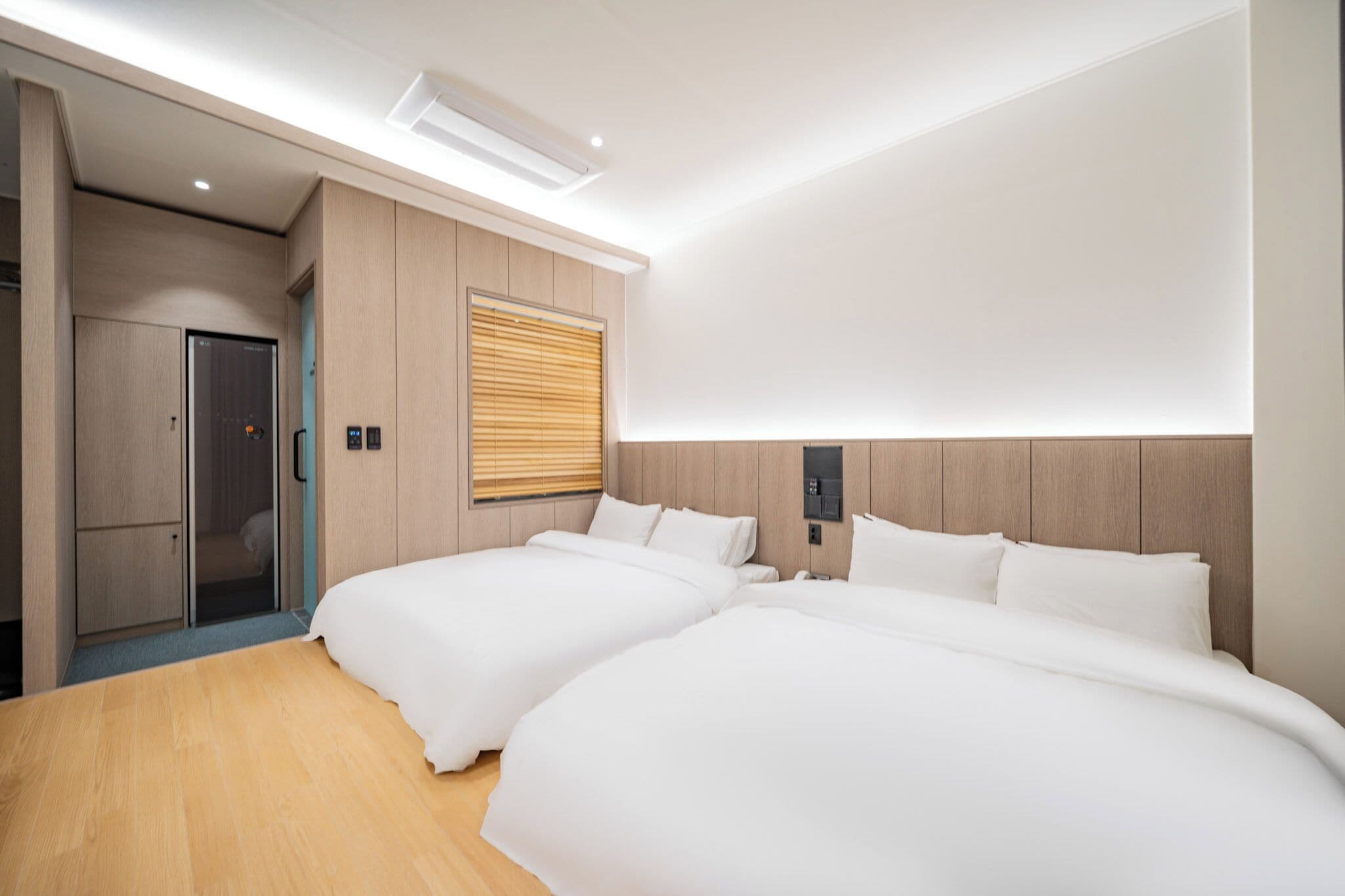 A modern hotel room with two beds, wooden flooring, a built-in wardrobe, and a large window with blinds.