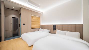 A modern hotel room with two beds, wooden flooring, a built-in wardrobe, and a large window with blinds.
