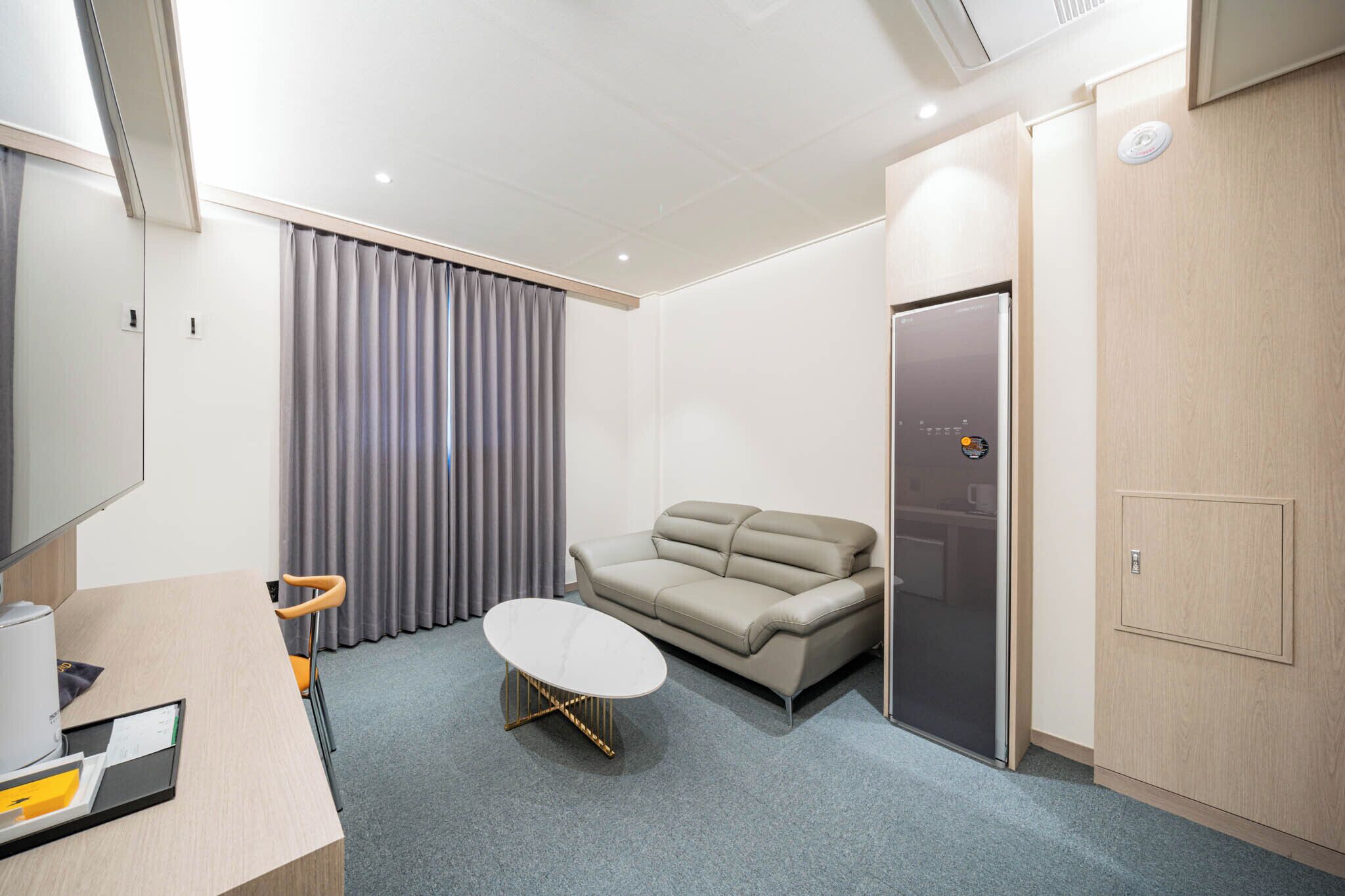 A modern hotel room with a sofa, a small table, a desk with a computer, and a door with a control panel.