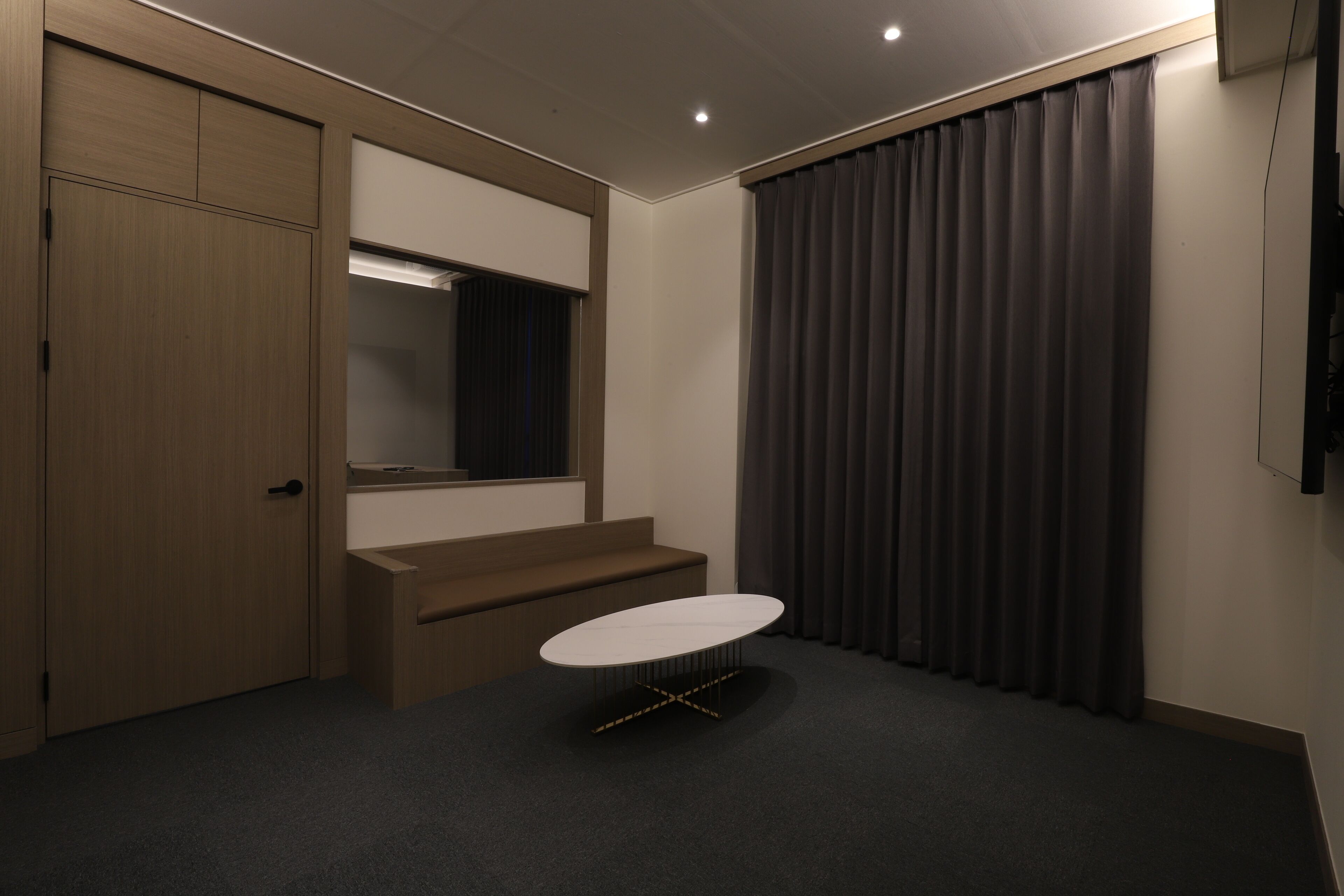 Ein Hotelzimmer mit Holztür, einem kleinen Tisch, einer Bank, einem großen Spiegel und einem dunklen Vorhang.