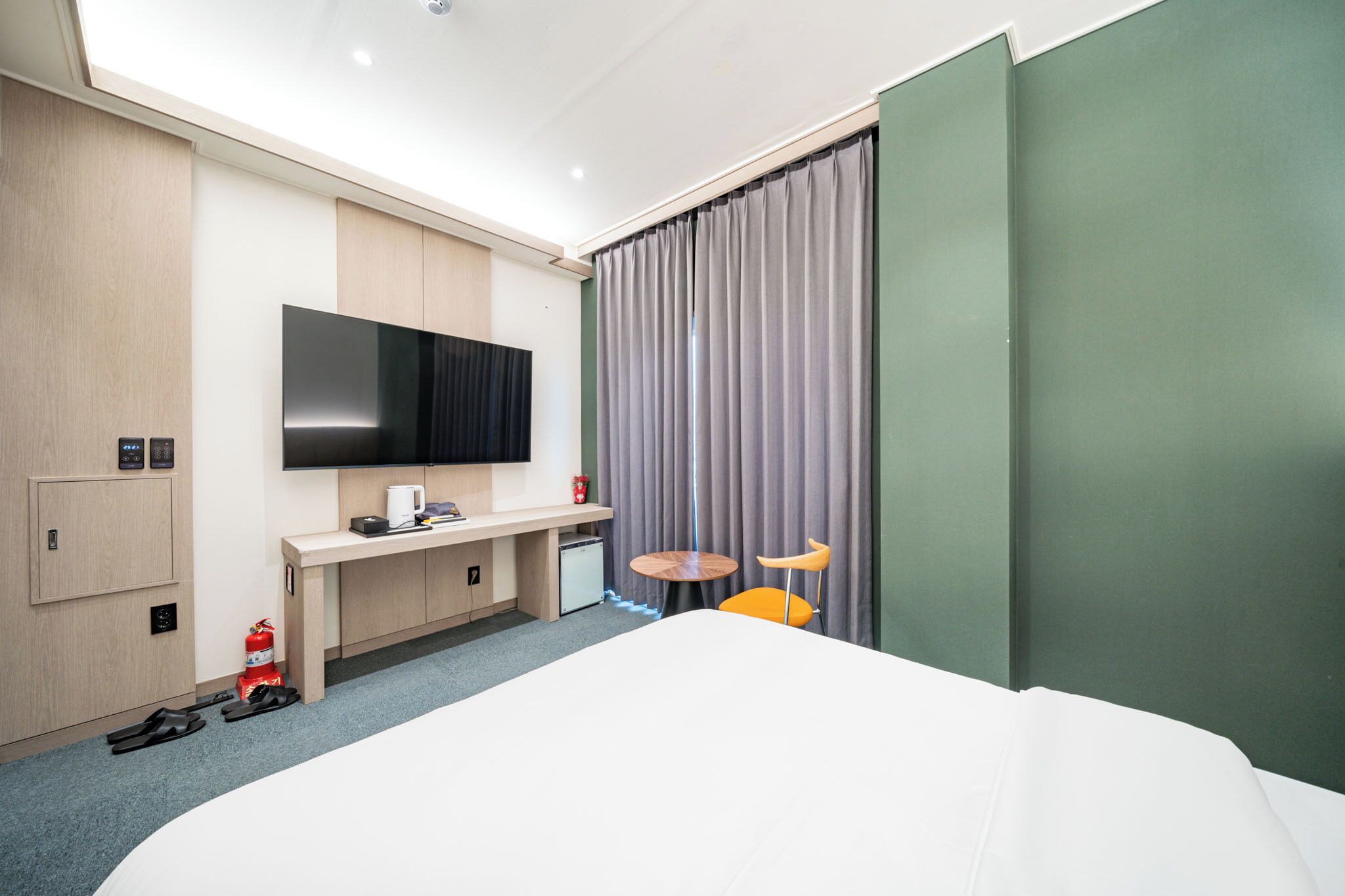 Camera d'albergo moderna con un letto grande, una televisione a schermo piatto montata a parete, una scrivania con una sedia, un tavolino rotondo con due sedie e un estintore sul pavimento.