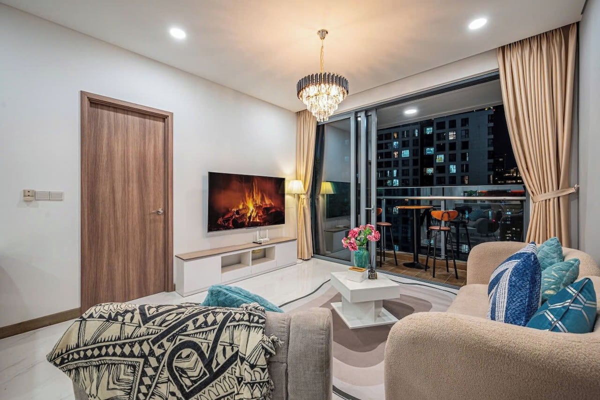 A modern living room with a flat-screen TV, a chandelier, a sofa, and a large window with curtains.