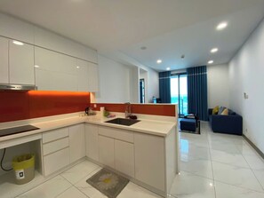 A modern kitchen with white cabinets, a built-in oven, and a sink. There is a dining area with a blue sofa and a window with blue curtains. - The Sunwah Pearl Residences By An Gia Hospitality (Ho Chi Minh City)