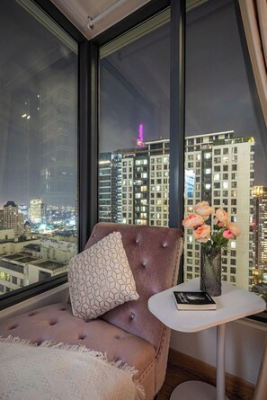 Ein modernes Zimmer mit großem Fenster, einer gepolsterten Sitzecke, einem kleinen Tisch mit Buch und Blumen sowie Blick auf die Stadt.