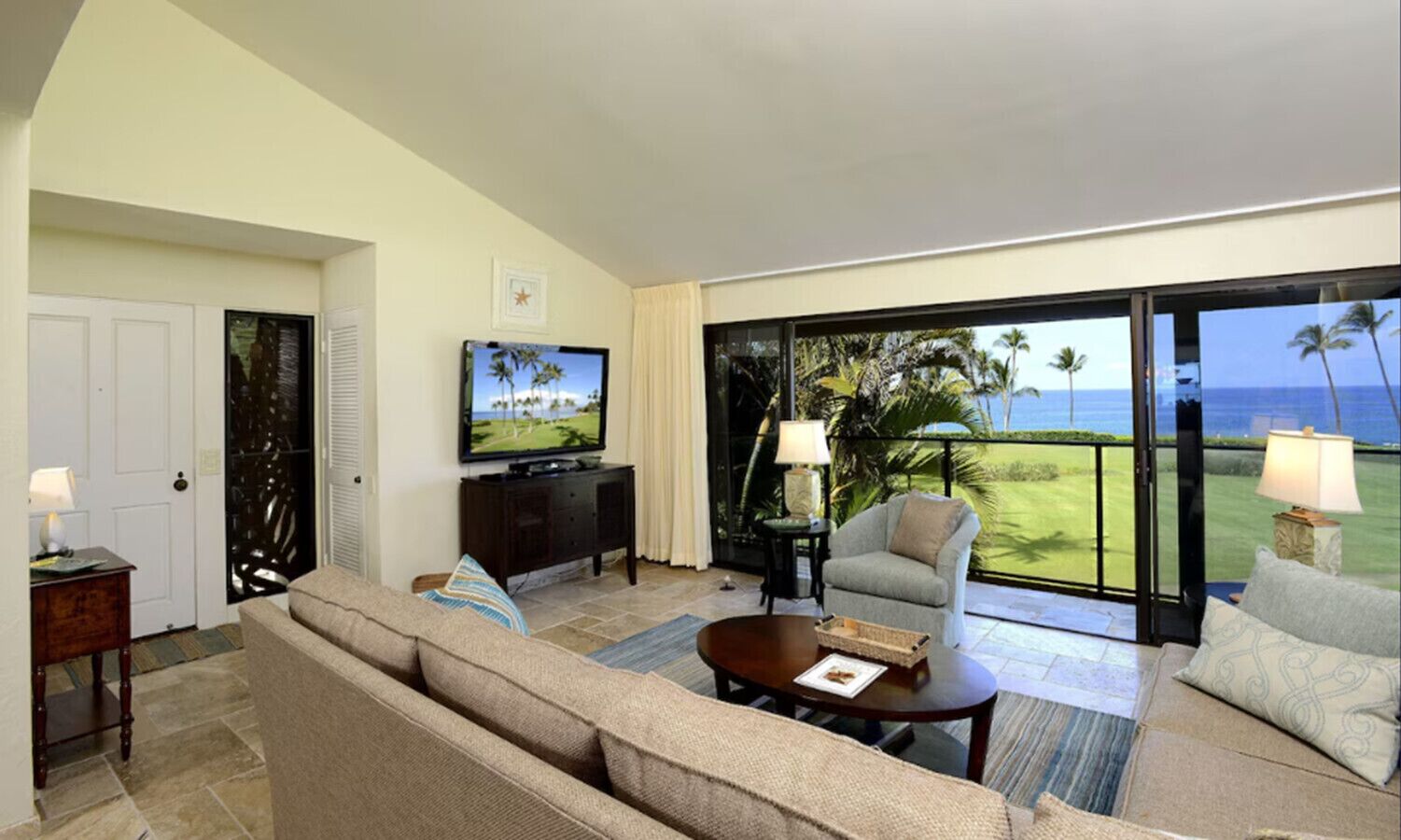 A spacious living room with a large flat-screen TV, comfortable seating, and a view of the ocean.