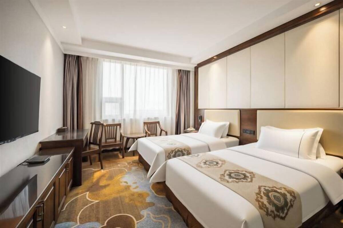 Habitación de hotel con dos camas, un escritorio con silla, televisor y un ventanal con cortinas.