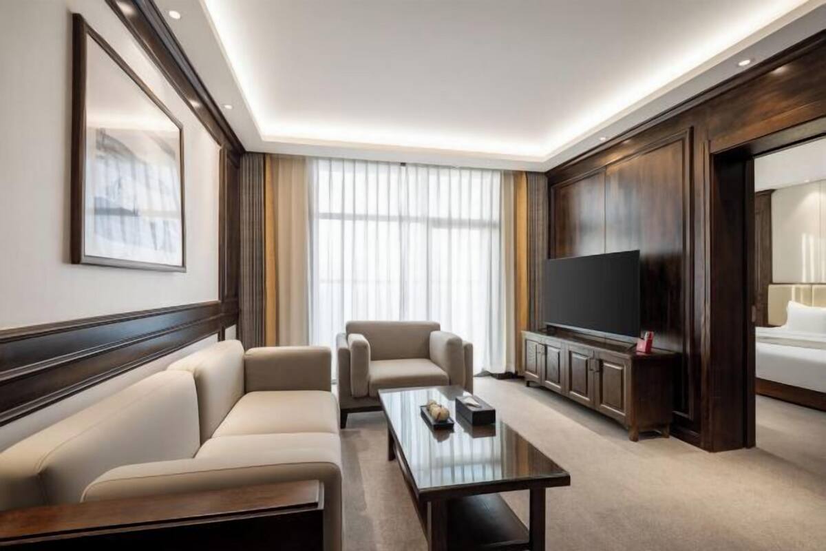 Habitación de hotel con sofá, mesa de centro, televisor de pantalla plana y ventana grande con cortinas.