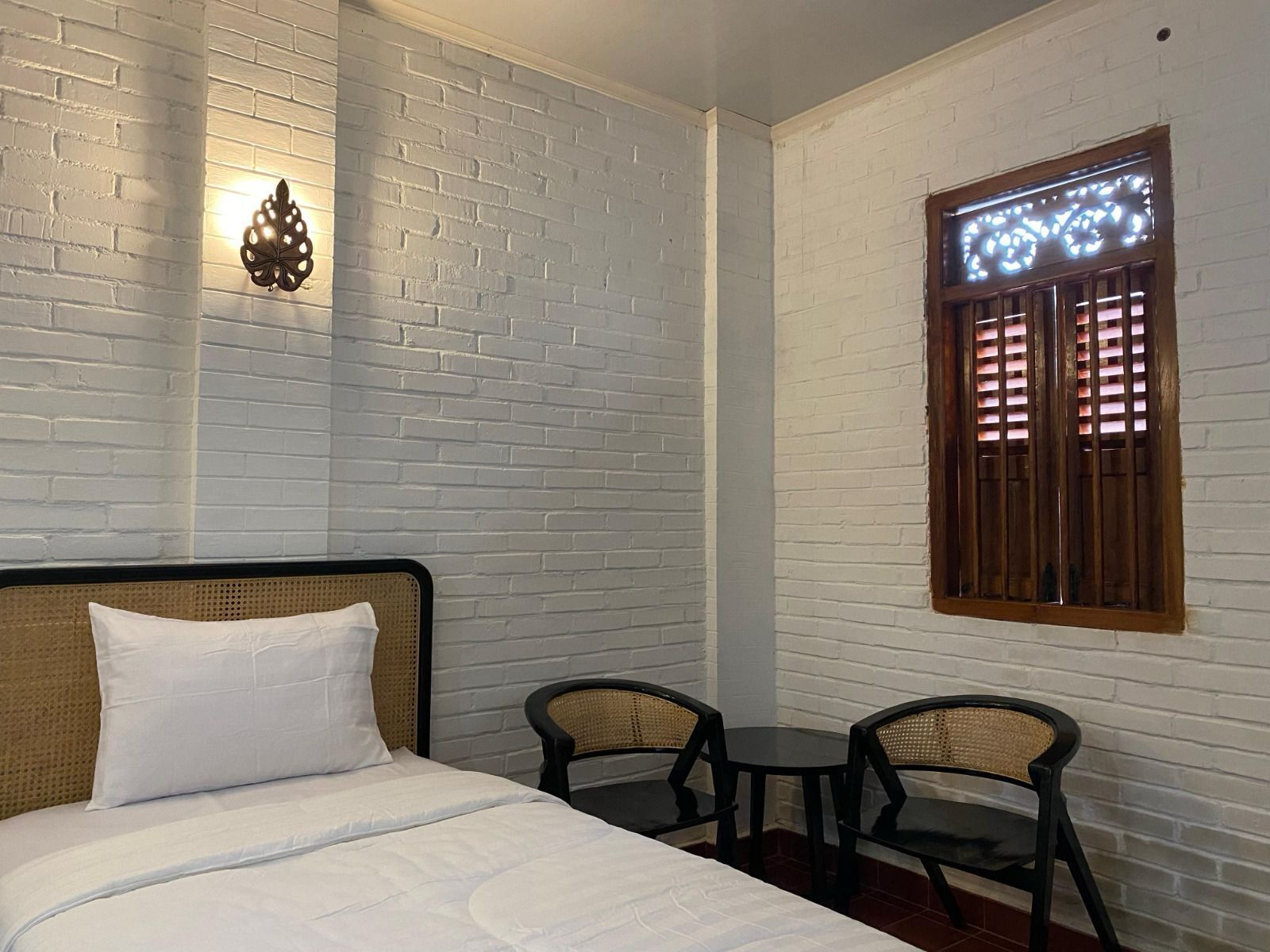 Una habitación con cama, dos sillas, una mesita pequeña, una lámpara decorativa en la pared y una ventana con marco de madera.