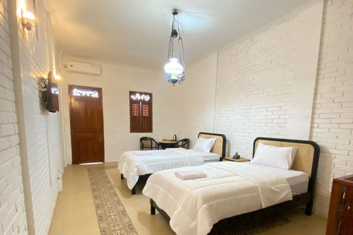 Habitació d'hotel amb dos llits, una catifa estampada, una làmpada penjant i una porta de fusta.
