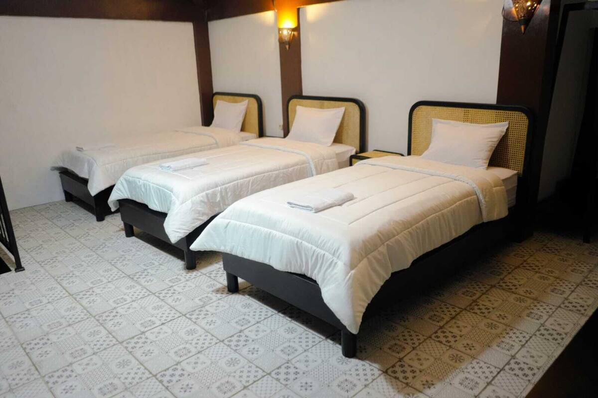 Habitació d'hotel amb tres llits individuals, cadascun amb un llençol blanc i una almoçada. La habitació té un paviment amb dibuixos i un lluminària fixat a la paret.