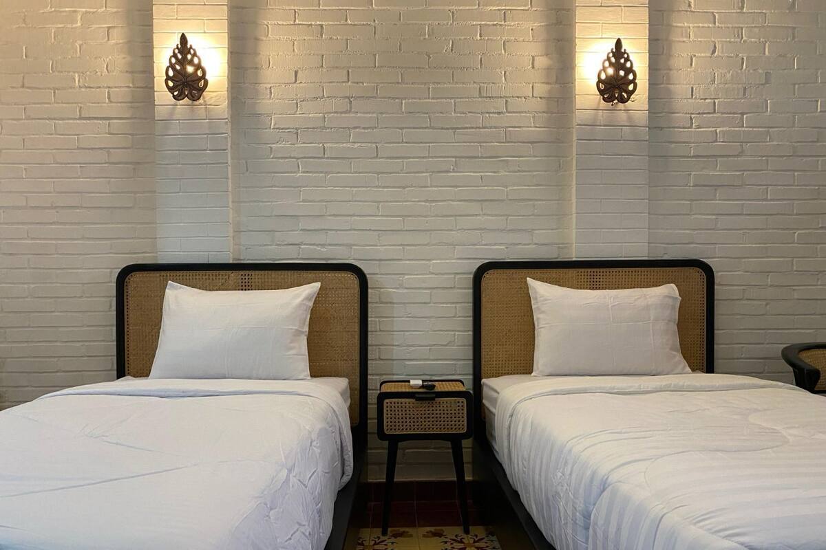 Habitació d'hotel amb dos llits individuals, roba de llit blanca, un tabler de palla i aplics de paret.