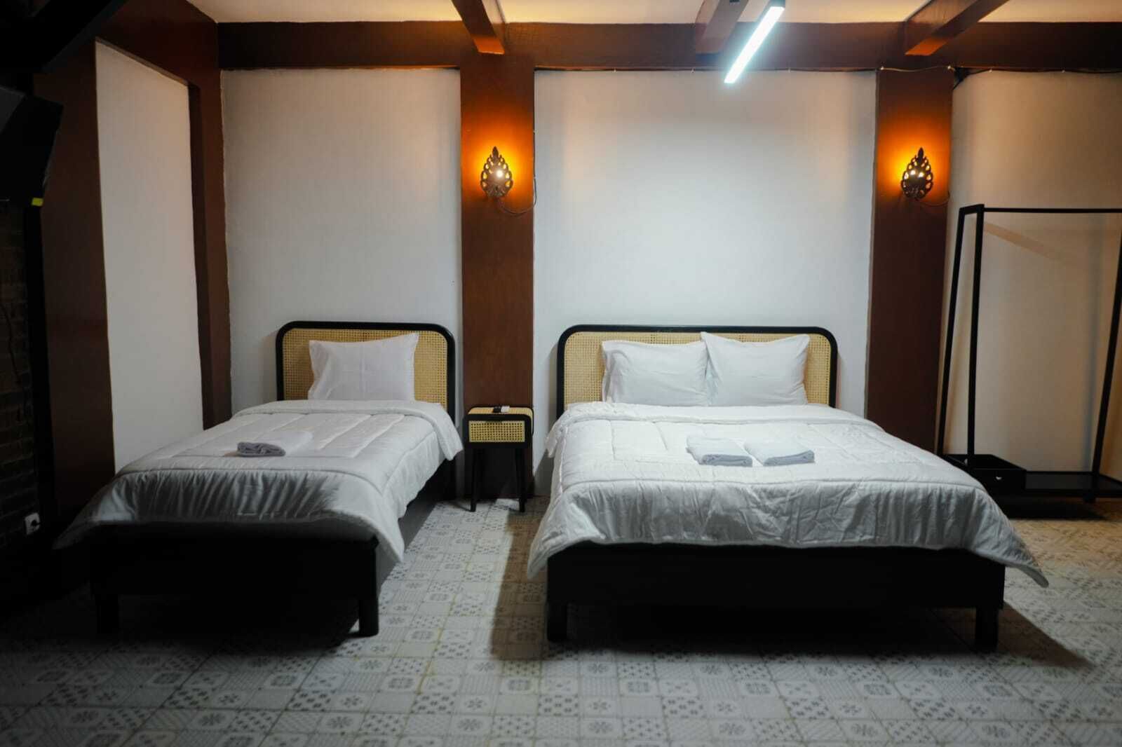 A hotel room with two single beds, each with a white bedspread and pillows, a small bedside table, and a wall-mounted light fixture.
