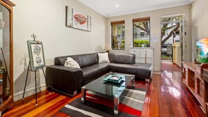 A living room with a leather sofa, a glass coffee table, and a red and white striped rug. There are two windows with blinds, a TV, and a cabinet.