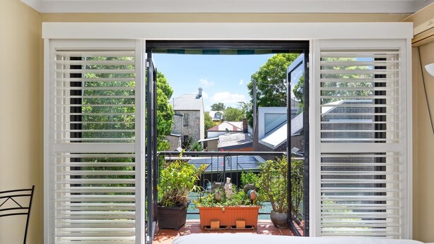 A bedroom with a large window, a bed, and a view of a garden and neighboring houses.