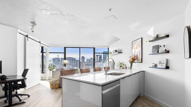 A modern kitchen with a large island, a dining area with a table and chairs, and a city view through large windows.
