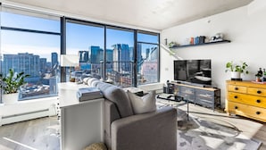 A modern living room with a large window offering a city view, a sofa, a TV, and a wooden dresser.