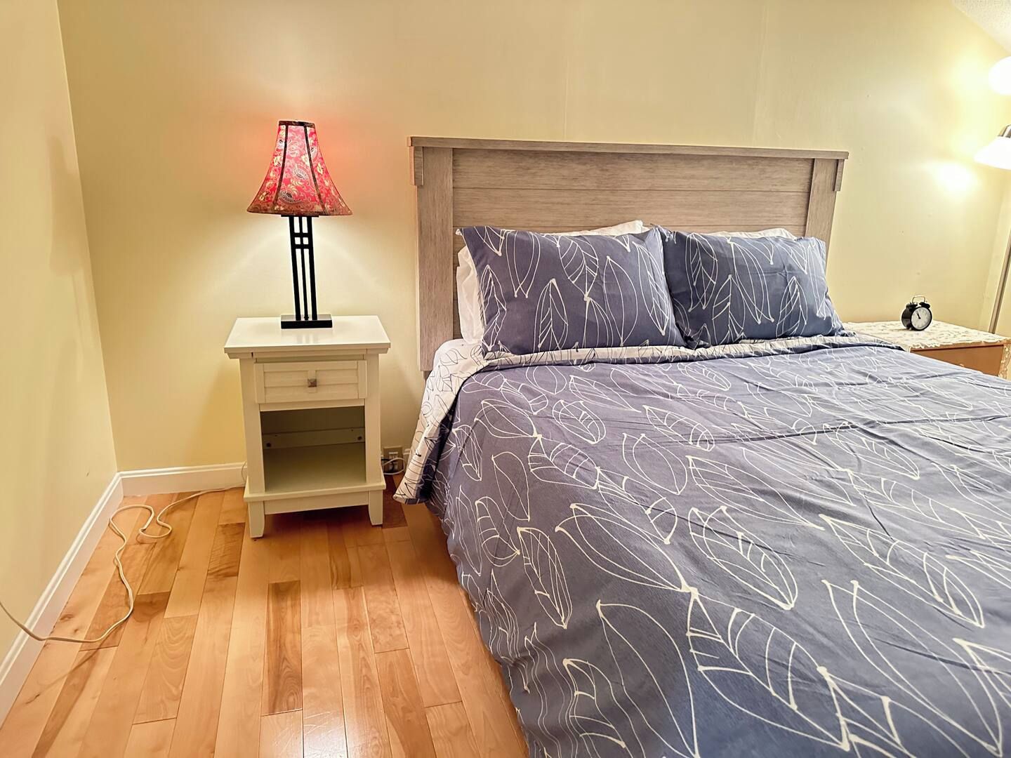 A bedroom with a bed, nightstand, lamp, and an alarm clock.