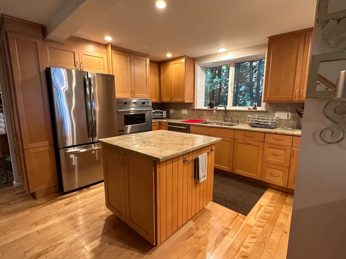 A kitchen with wooden cabinets, a stainless steel refrigerator, an oven, and a window with a view of trees.