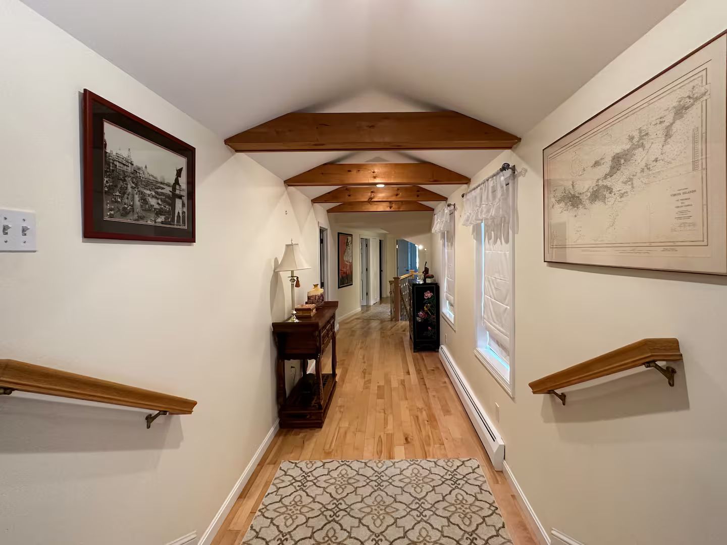 A hallway with wooden beams, a patterned rug, a desk with a lamp, and a framed map on the wall.