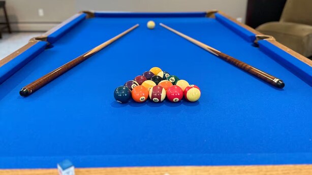 A pool table with a blue felt surface, surrounded by wooden railings and a window with a plaid curtain. - Waterfront Oasis~Pool Table~Deck~Dock~Garage (Dover)