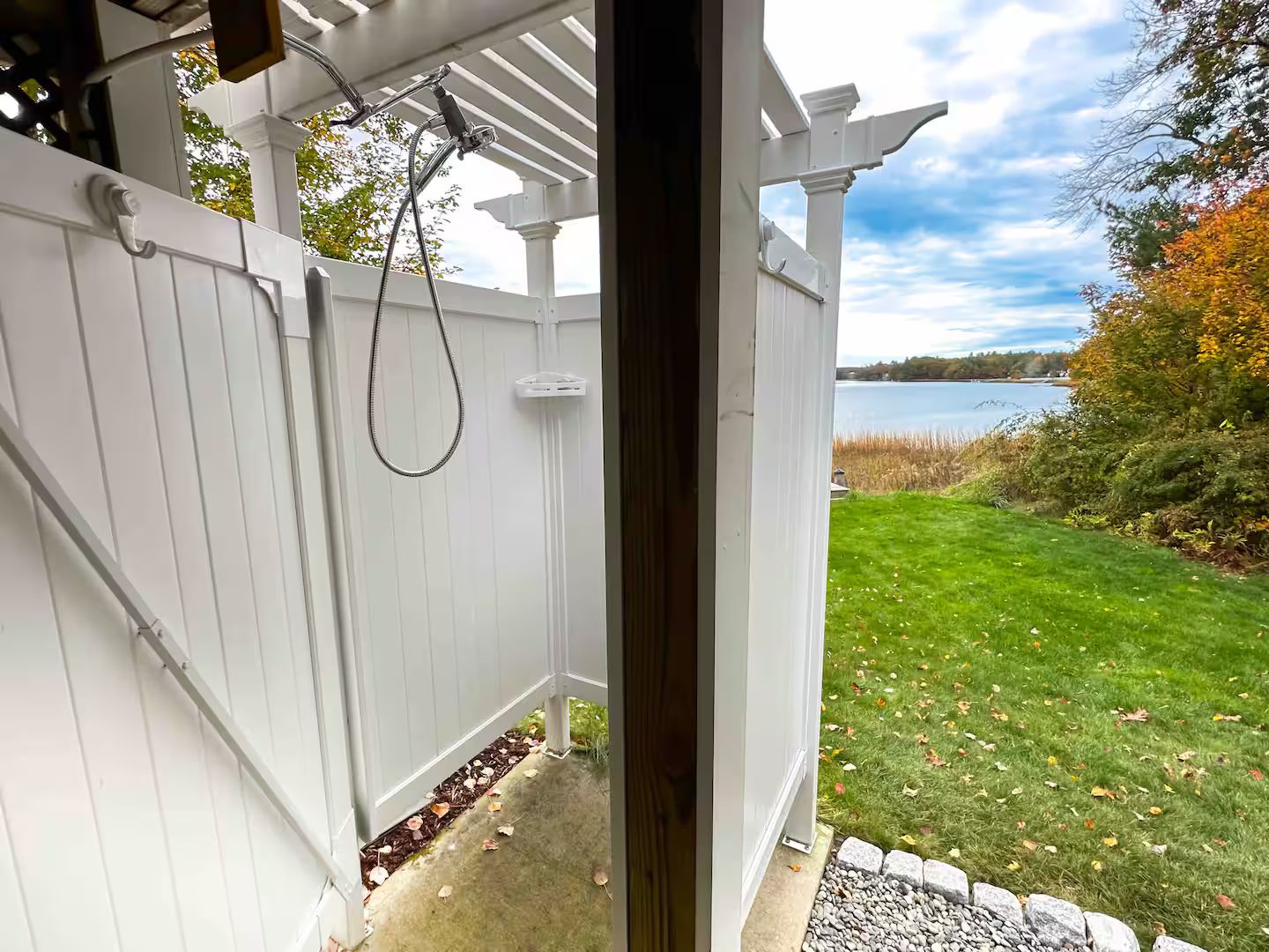 A white wooden pergola with a hose attached, overlooking a grassy yard with a body of water in the background.