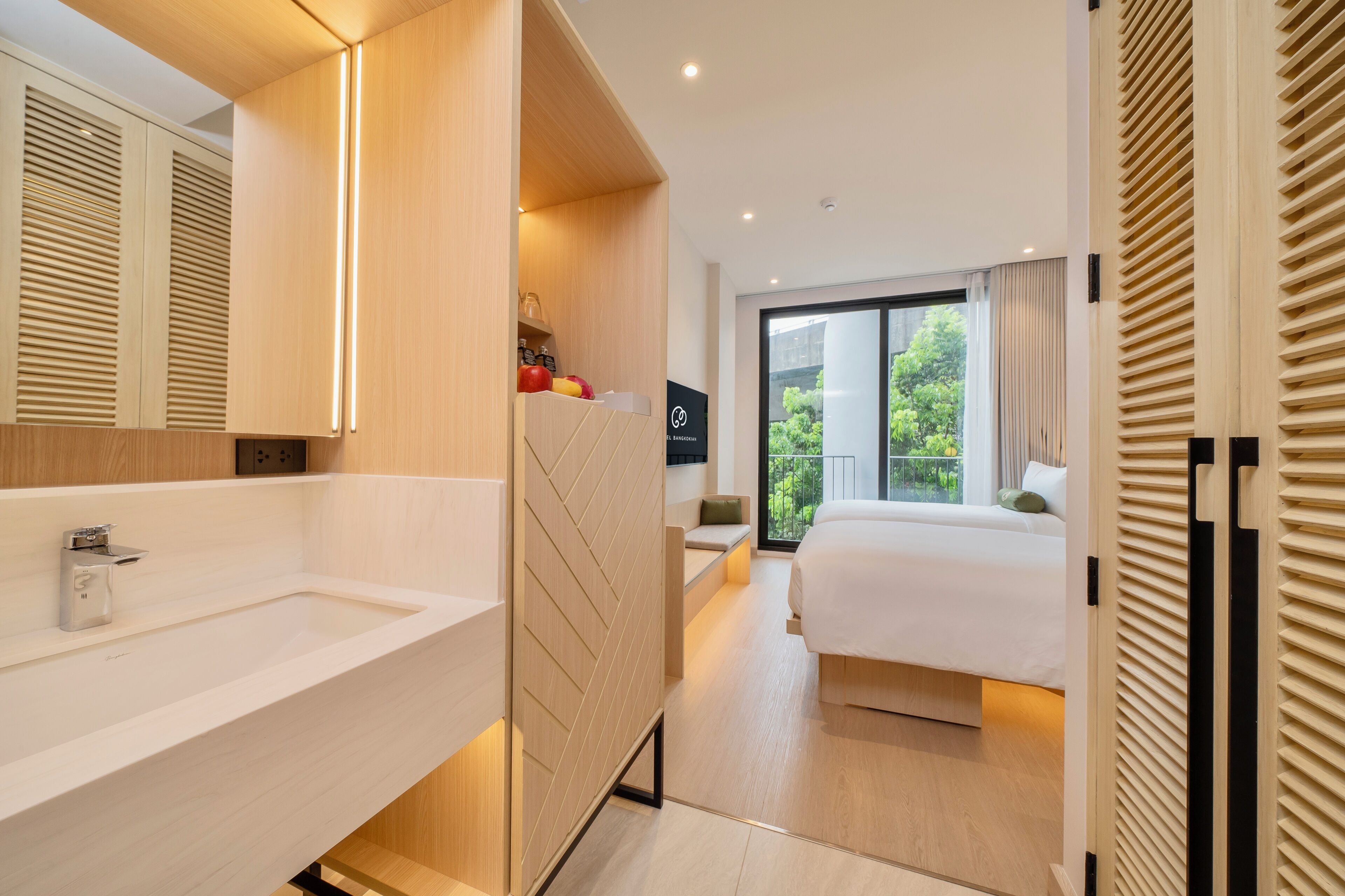 Una habitación de hotel moderna con una cama grande, un baño con lavamanos y espejo, y un balcón con vistas a zonas verdes.