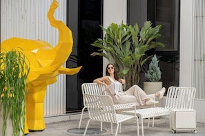 A person relaxing on a white metal chair in an outdoor seating area with a yellow abstract sculpture and potted plants. - Hotel Bangkokian (Bangkok)
