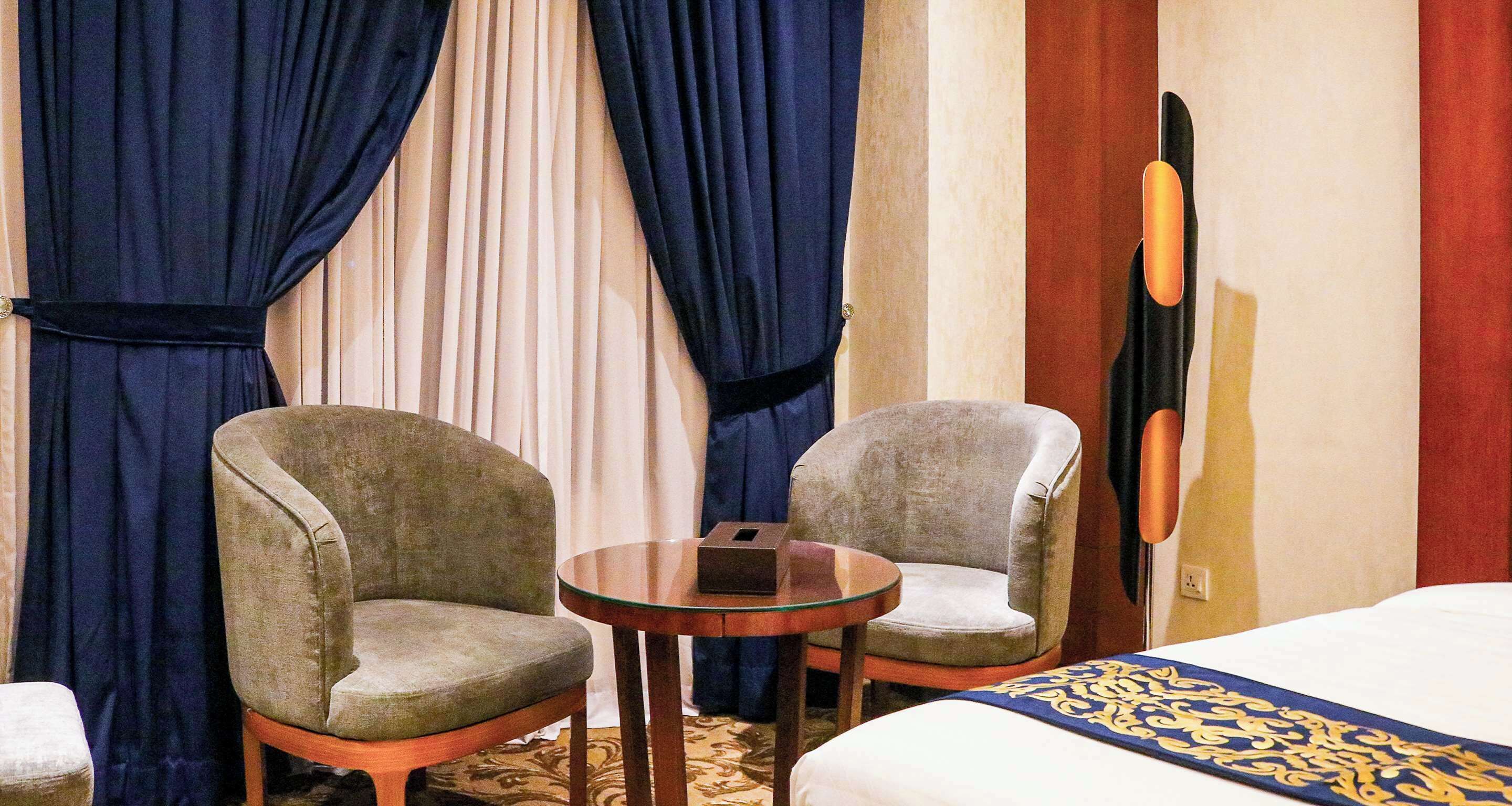 Habitación de hotel con dos sillones, una mesita redonda, una cama con colcha estampada y un cuadro grande en la pared.
