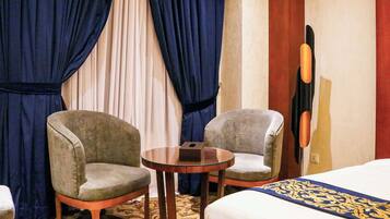 Habitación de hotel con dos sillones, una mesita redonda, una cama con colcha estampada y un cuadro grande en la pared.
