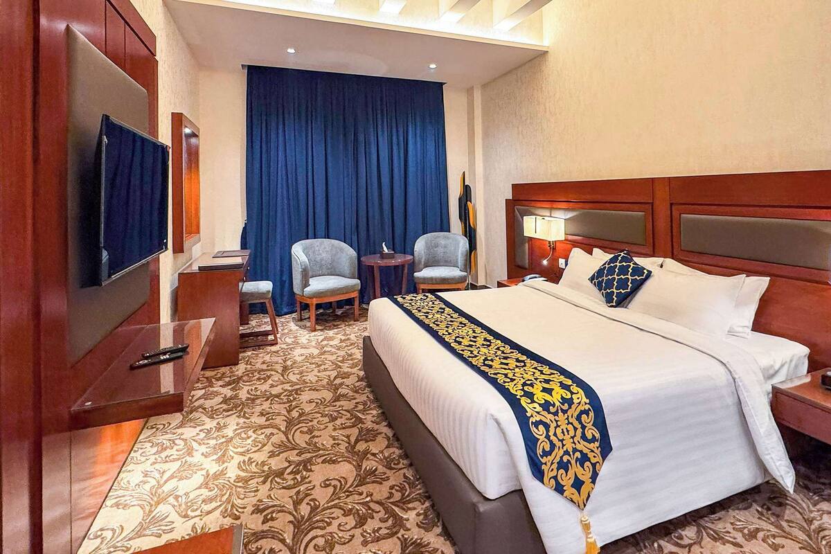 A hotel room with a large bed, two armchairs, a small table, a TV, and a desk with a lamp.