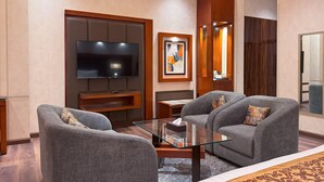Una habitación de hotel moderna con un televisor de pantalla plana, dos sillones grises, una mesa de centro de vidrio, un mueble de entretenimiento de madera y un espejo grande.