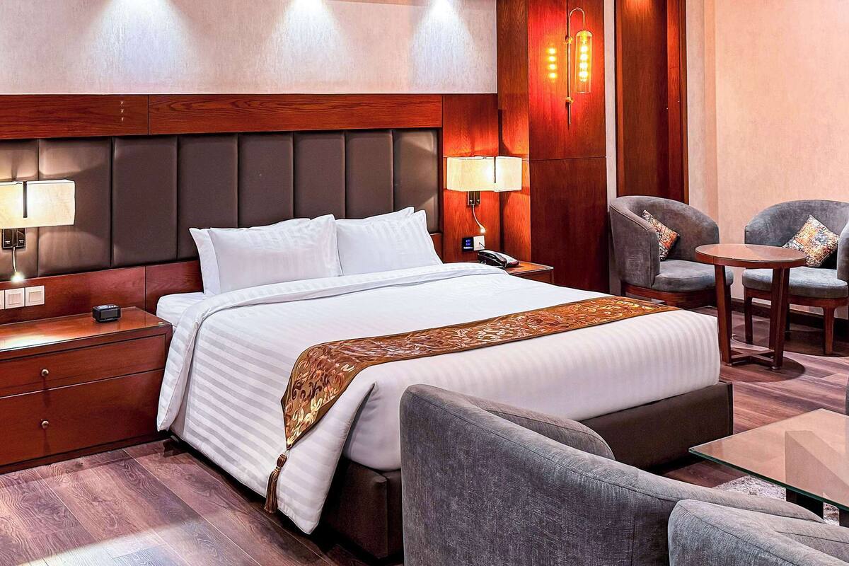 A hotel room with a large bed, two armchairs, a small table, and a nightstand with a lamp.