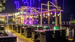 Un área de comedor al aire libre con mobiliario de mimbre y mesa de billar, rodeada por una barandilla y guirnaldas de luces.