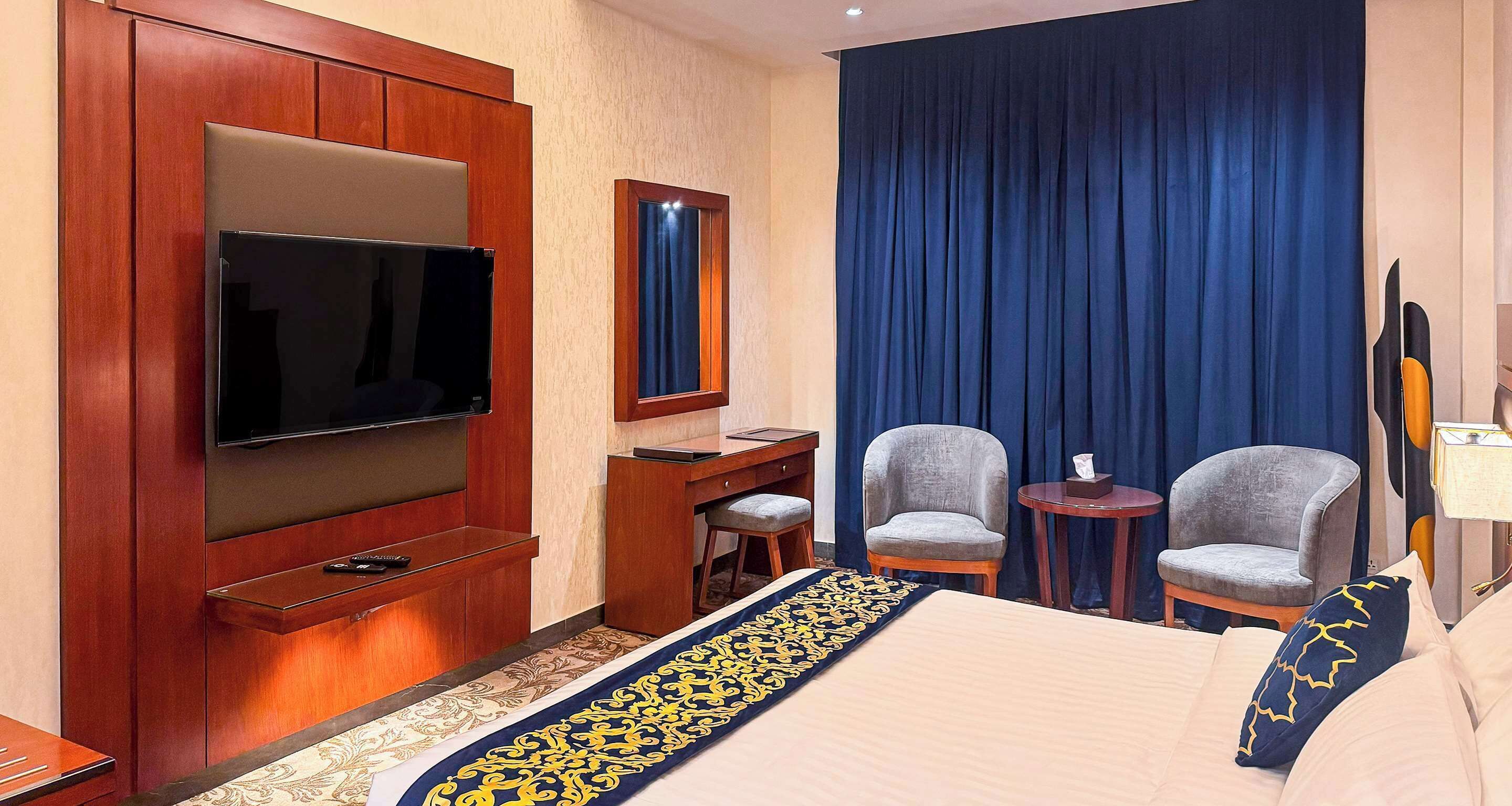 Habitación de hotel con cama, televisión, escritorio, dos sillones y un ventanal con cortinas.