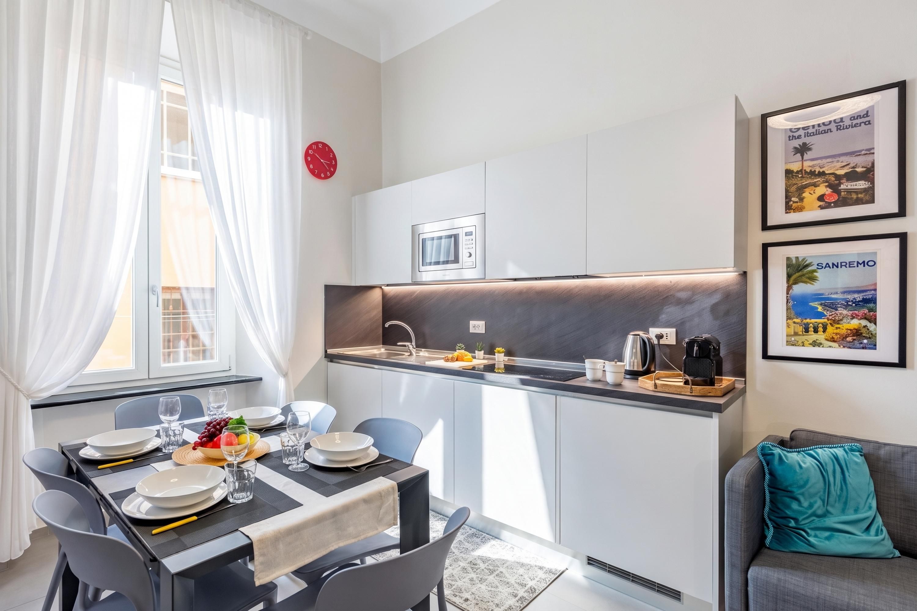 A modern kitchen with a built-in microwave, a sink, and a coffee machine. There is a dining area with a table set for two, and a sofa in the corner.