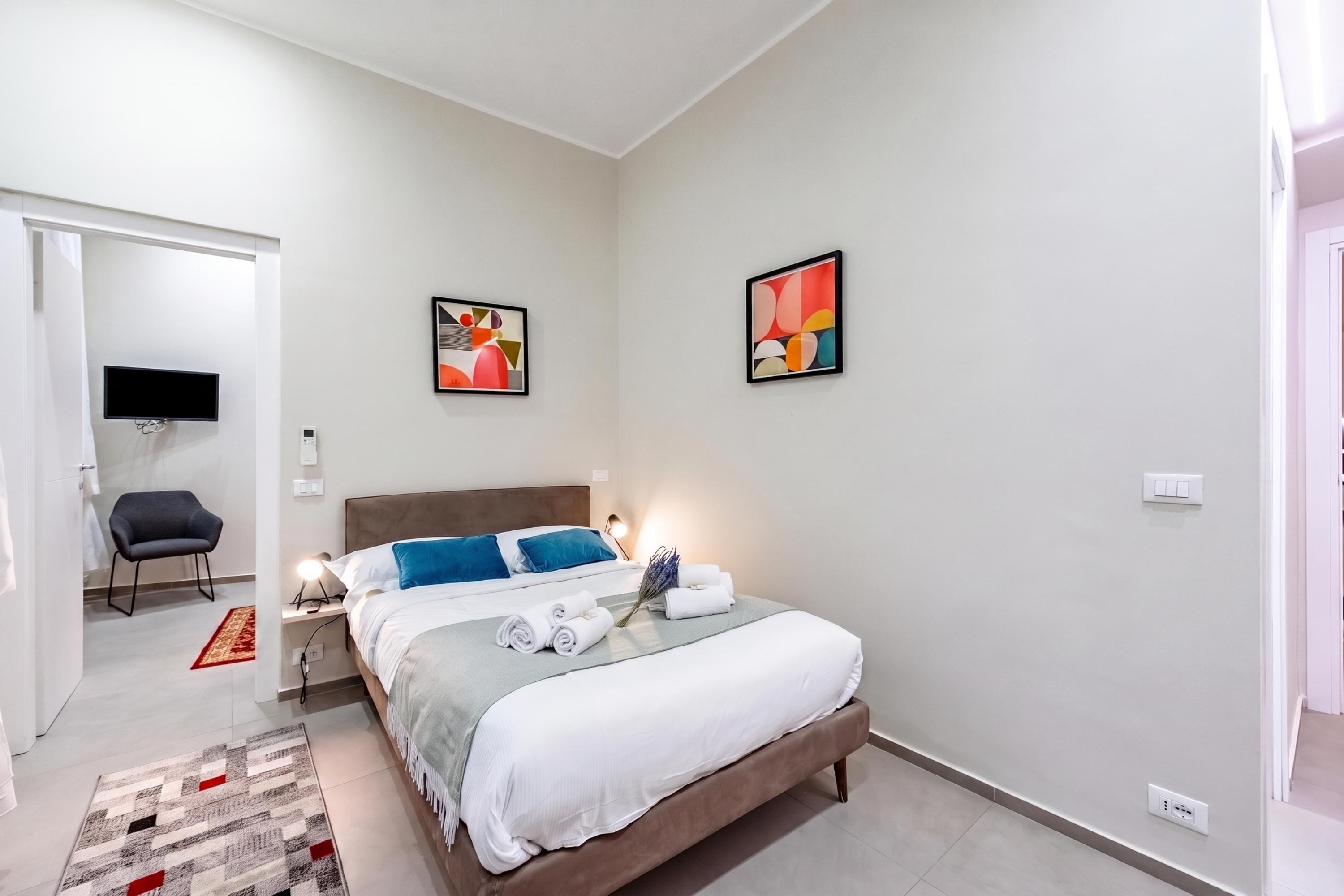 A modern bedroom with a bed, a desk with a chair, a flat-screen TV, and artwork on the walls.