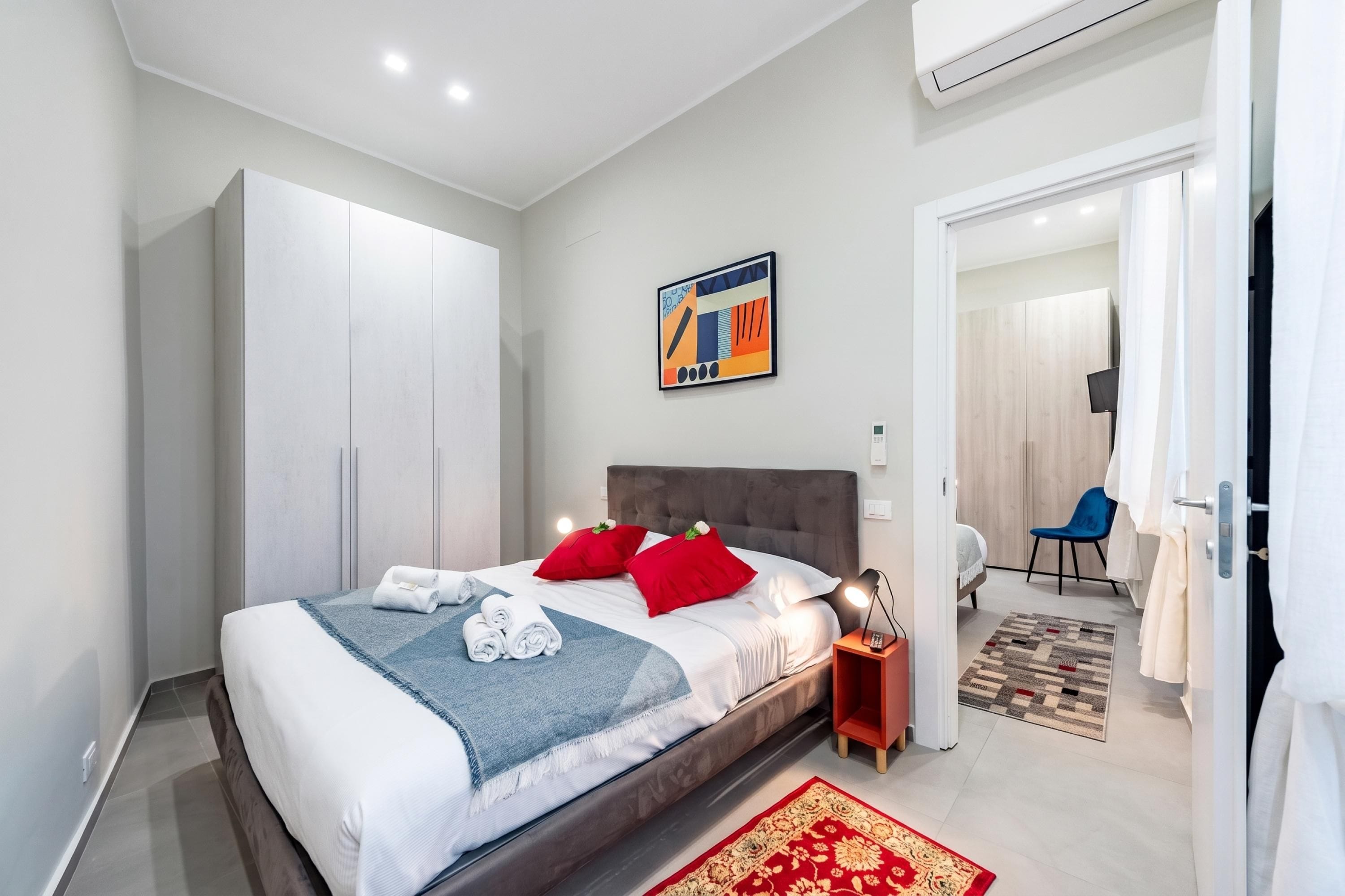 A modern hotel room with a bed, a nightstand, a painting, a wardrobe, and a view into the adjacent room.