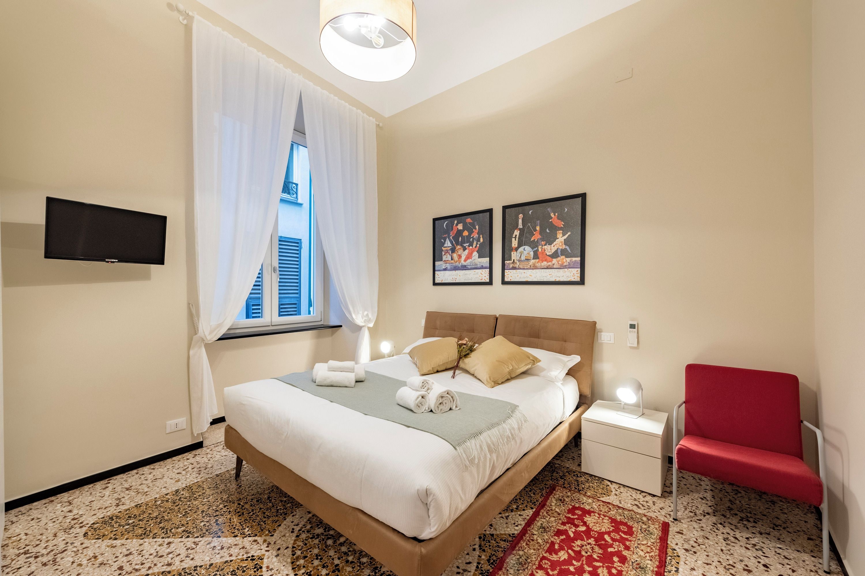 A neatly arranged bedroom with a bed, bedside tables, a TV, artwork, and a window with curtains.