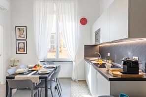 Une cuisine moderne dotée d’un four encastré et d’un évier, un coin repas avec une table dressée pour deux personnes, et une fenêtre ornée de rideaux légers.