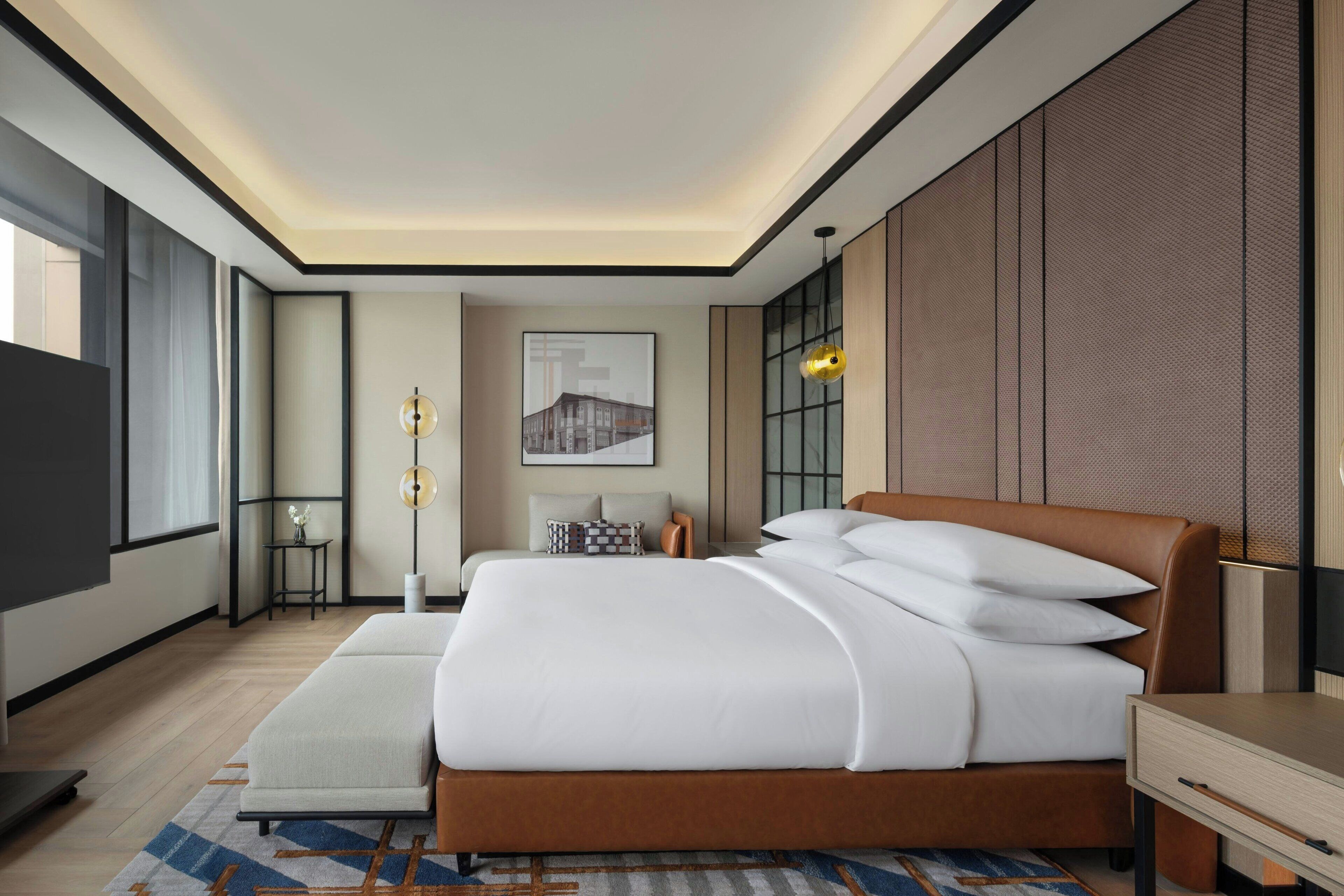 A modern hotel room with a large bed, a bedside table, a nightstand, a floor lamp, and a framed picture on the wall.